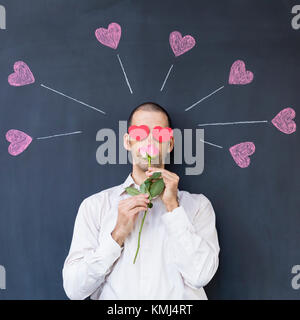 Seul adulte homme blanc avec des yeux en forme de coeur portant une chemise blanche et debout devant un tableau noir avec coeurs peints tenant une rose. Banque D'Images