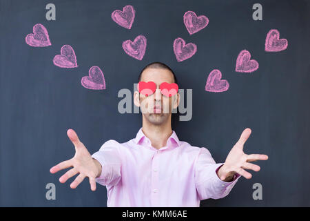 Seul adulte homme blanc avec des yeux en forme de coeur portant un chandail rose et debout devant un tableau noir avec coeurs peints en tendant ses mains. Banque D'Images