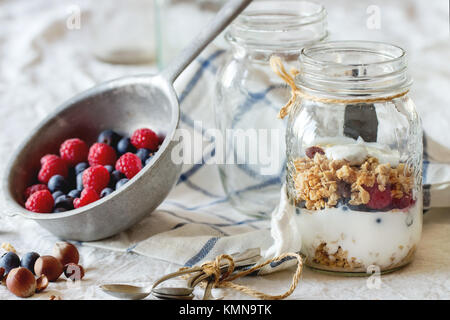 Bocal en verre avec du muesli et yaourt servi avec des noix, des framboises et des mûres dans une passoire sur textile blanc vintage dans la lumière du jour Banque D'Images