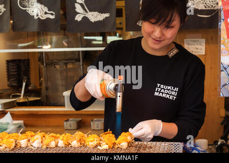 Marché aux poissons de Tsukiji, Tokyo, Japon. - 7 août 2023 : femme asiatique japonaise griller et faire cuire des noix de Saint-Jacques et des œufs de mer ​​urchin brochette sur la nourriture de rue japonaise Banque D'Images