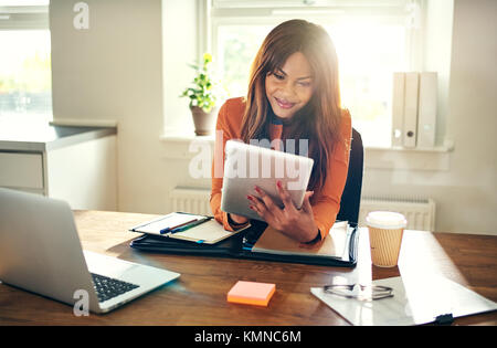 Souriante jeune femme entrepreneur africain travaillant en ligne avec une tablette numérique alors qu'assis à un bureau dans son bureau à domicile Banque D'Images