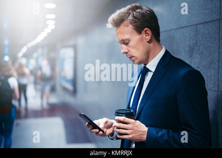 Jeune homme de boire du café et de la lecture des messages texte sur son téléphone cellulaire alors qu'il se trouvait sur une plate-forme du métro durant son trajet du matin Banque D'Images