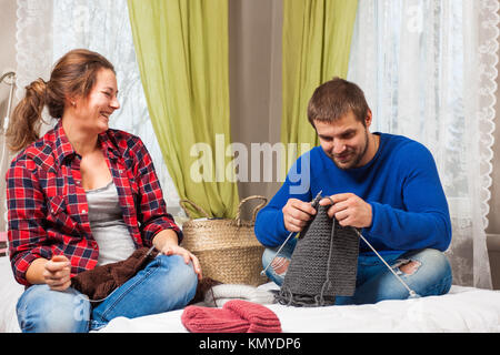 Jeune femme enceinte sourit et enseigne le jeune homme en pull bleu et jeans comment tricoter avec des aiguilles à tricoter à partir de fils naturels chandail gris assis Banque D'Images