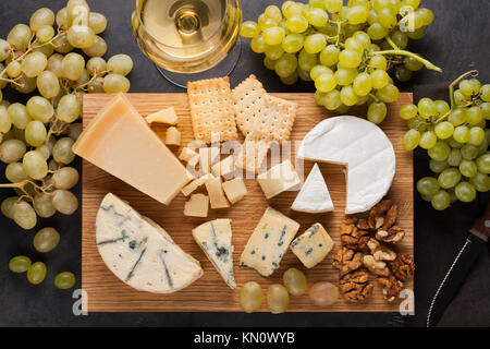 Assortiment de fromages, des raisins blancs, noix, craquelins et vin blanc sur une planche de bois. Nourriture pour une date romantique sur une pierre sombre arrière-plan. Vue d'en haut. Banque D'Images