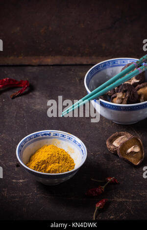 Curcuma poudre et les champignons shiitake dans des bols en porcelaine, servi avec Red Hot Chili Peppers et baguettes turquoise foncé sur table. Voir la série Banque D'Images