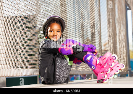 Portrait de belle fille africaine portant des patins à roulettes et les équipements de protection, assis à côté de la clôture de grillage métallique à une rollerdrom Banque D'Images