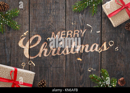 Texte de vœux de Noël à la main sur la surface en bois sombre avec des décorations de Noël. Les cadeaux, les branches de sapin et de cocottes à côté. Banque D'Images