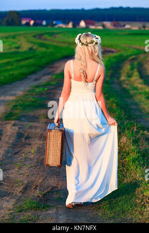 Fille blonde wearing white Robe longue de style rétro avec valise sur campagne environnante Banque D'Images