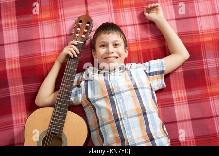 Garçon jouant de la musique à la guitare, se trouve sur une couverture à carreaux rouge, vue du dessus Banque D'Images