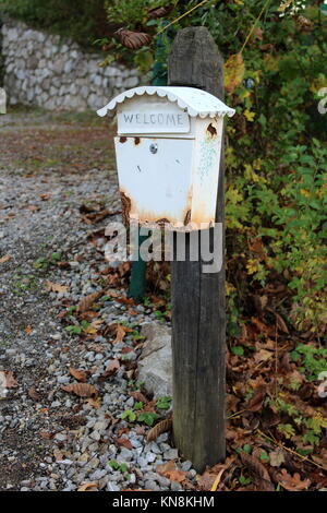 Boîte aux lettres blanc rouillé avec panneau de bienvenue monté sur poteau en bois ancien à côté entrée couverte de gravier avec des feuilles vertes et brunes en arrière-plan Banque D'Images