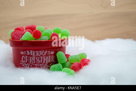 Joyeux Noël rouge bol rempli de gomme rouge et vert avec quelques gouttes de débordement dans 'Snow' contre un mur en bois Banque D'Images