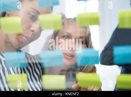 Two businesswomen derrière mur de verre pointant notes adhésives Banque D'Images