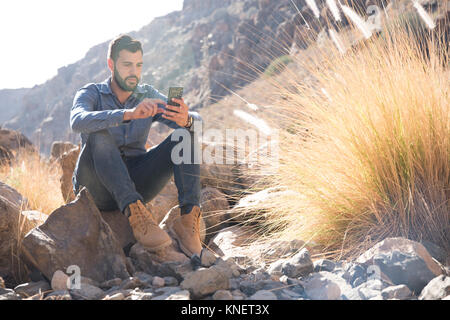 Young male hiker assis sur boulder looking at smartphone en vallée, Las Palmas, Canaries, Espagne Banque D'Images