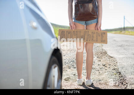 Jeune femme debout à côté de voiture, holding de l'autostop panneau disant 'anywhere', low section Banque D'Images