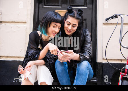 Deux jeunes femmes élégantes amis assis porte looking at smartphone Banque D'Images