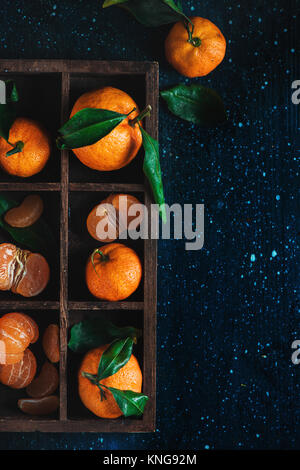 Mandarines dans une caisse de bois sur un fond sombre. Un assortiment de clémentines avec des feuilles vertes. La photographie alimentaire sombre avec des fruits orange vif et Banque D'Images