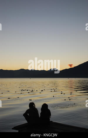 Un couple s'asseoir sur une jetée au lever du soleil dans le lac Atitlan au Guatemala qui est entouré de hautes falaises et dominé par trois volcans Banque D'Images