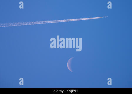 Contrails sur la lune. Avion de ligne à réaction en hauteur créant des traînées de vapeur devant un croissant de lune déclinant dans un ciel bleu jour. Voler haut Banque D'Images