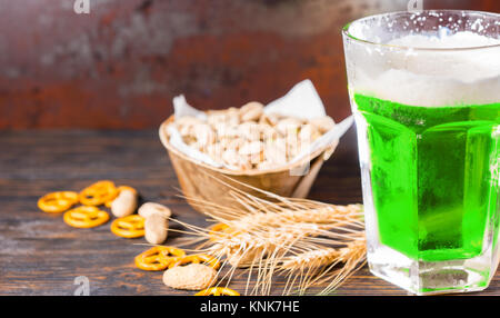 Verre à bière verte et une tête de mousse près de la plaque avec les pistaches, le blé, les arachides et les bretzels petits éparpillés sur dark 24. Aliments et boissons conc Banque D'Images