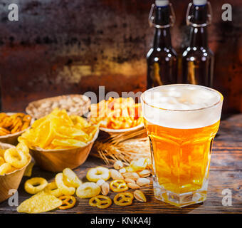 Les plaques avec des collations près de deux bouteilles et un verre de bière, de blé, d'écrous et les bretzels dispersés sur un bureau en bois sombre. Aliments et boissons concept Banque D'Images