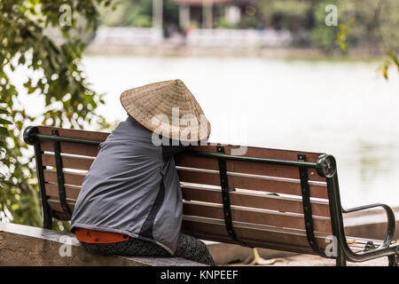 Femme avec chapeau de bambou vietnamien typique se reposant sur un banc au bord du lac Banque D'Images