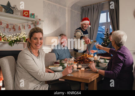 Mature femme sourit à l'appareil photo tout en appréciant un verre de vin au dîner de Noël de sa famille. Banque D'Images