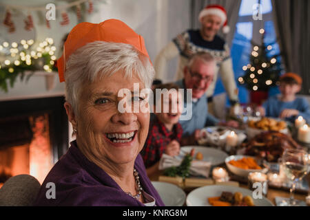 Heureux grand-mère à un dîner de Noël en famille. Elle sourit à l'appareil photo tout en portant un chapeau de fête de Noël. Banque D'Images