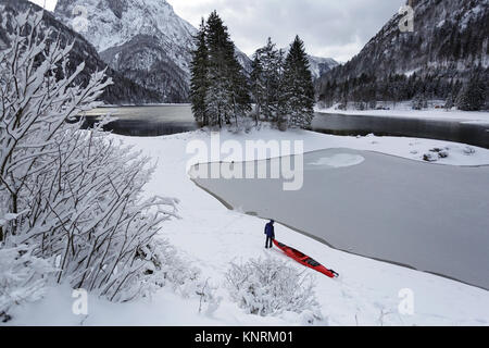 Homme portant un kayak rouge sur une froide journée d'hiver à l'lakeLago congelés en partie di Predil d'avoir certaines activités récréatives d'hiver dans la neige, les Alpes, l'Italie. Banque D'Images