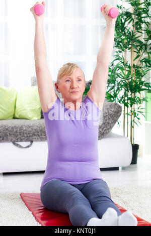 Hauts femme assise sur un tapis à la maison et de l'exercice avec des poids Banque D'Images