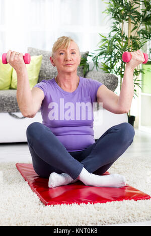 Hauts femme assise sur un tapis à la maison et de l'exercice avec des poids Banque D'Images
