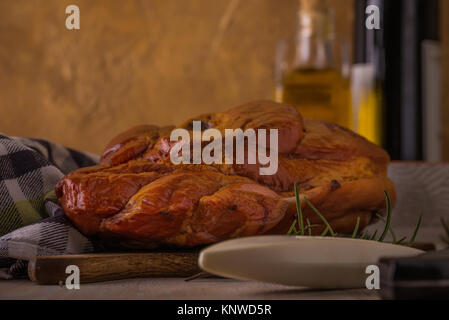 Photo horizontale avec de gros morceau de viande de porc fumé avec de l'os placé sur une planche à découper. Serviette bleu, broyeur, fourchette, bols et romarin sont autour avec w Banque D'Images