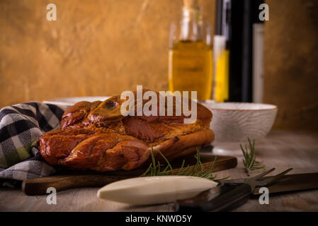 Photo horizontale avec de gros morceau de viande de porc fumé avec de l'os placé sur une planche à découper. Serviette bleu, broyeur, fourchette, bols et romarin sont autour avec w Banque D'Images