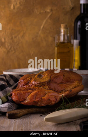 Photo verticale avec de gros morceau de viande de porc fumé avec de l'os placé sur une planche à découper. Serviette bleu, broyeur, fourchette, bols et romarin sont autour avec win Banque D'Images