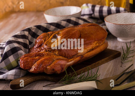 Photo horizontale avec de gros morceau de viande de porc fumé avec de l'os placé sur une planche à découper. Serviette bleu, broyeur, fourchette, bols et romarin sont autour avec w Banque D'Images