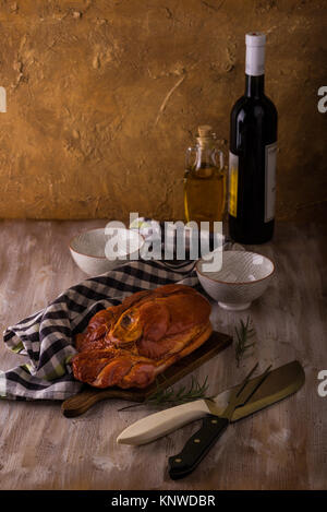 Photo verticale avec de gros morceau de viande de porc fumé avec de l'os placé sur une planche à découper. Serviette bleu, broyeur, fourchette, bols et romarin sont autour avec win Banque D'Images