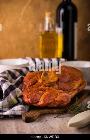 Photo verticale avec de gros morceau de viande de porc fumé avec de l'os placé sur une planche à découper. Serviette bleu, broyeur, fourchette, bols et romarin sont autour avec win Banque D'Images