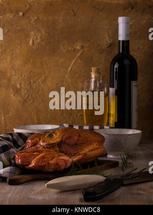 Photo verticale avec de gros morceau de viande de porc fumé avec de l'os placé sur une planche à découper. Serviette bleu, broyeur, fourchette, bols et romarin sont autour avec win Banque D'Images