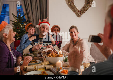 Family having Christmas dinner sont ensemble. Le grand-père est à l'aide d'un smartphone pour prendre une photo ridicule de sa famille. Banque D'Images