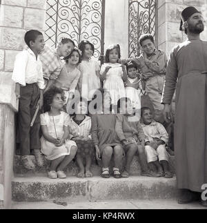 Années 1950, tableau historique, un homme barbu prêtre avec chapeau et la robe a un peu de plaisir avec un groupe d'enfants assis et debout sur les marches à l'extérieur d'une entrée fermée, la Jordanie. Banque D'Images