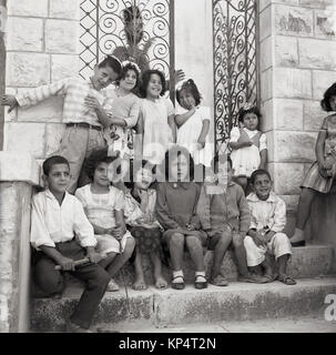 Années 1950, tableau historique, un homme barbu prêtre orthodoxe chrétienne a de l'amusement se cacher derrière certaines portes comme un groupe d'enfants s'asseoir et se tenir sur les marches et sourire et rire pour une photographie, la Jordanie. Banque D'Images