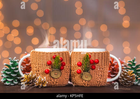 Deux tasses fumante blanc fait main en tricot mignon brun couvre avec des décorations de noël fête des Lumières floues en arrière-plan d'orange garland Banque D'Images