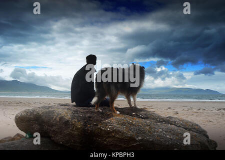 Homme avec rough collie, assis sur le rocher sur la plage à Dingle, Irlande. Collines en distance et dramatique ciel au-dessus. Banque D'Images