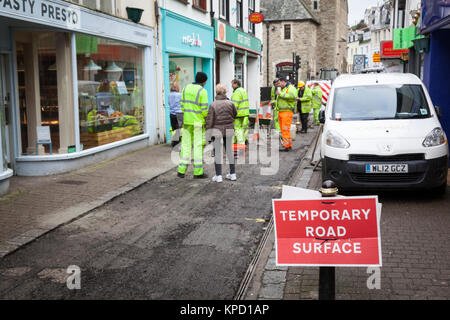 Un panneau rouge avertit d'une surface de la route comme travailleurs temporaires en jaune vif hi-viz jackets préparer une nouvelle surface d'une route étroite à travers le centre de Banque D'Images