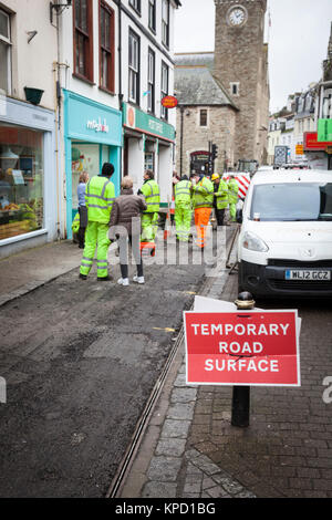 Un panneau rouge avertit d'une surface de la route comme travailleurs temporaires en jaune vif hi-viz jackets préparer une nouvelle surface d'une route étroite à travers le centre de Banque D'Images