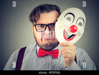 Jeune homme bouleversé de froncer tout en regardant la caméra et holding happy clown masque. Banque D'Images