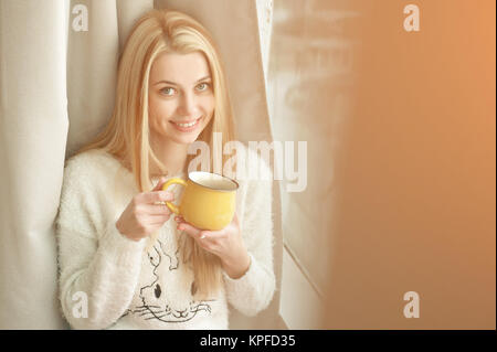Jeune femme debout près de la fenêtre avec une tasse de café, à l'extérieur et souriant. Lumière naturelle en plein air. Banque D'Images