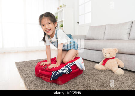 Happy petit enfant fille agenouillée sur valise face à huis clos et en appuyant sur la fermeture d'essayer de trop plein voyage à la maison. Banque D'Images