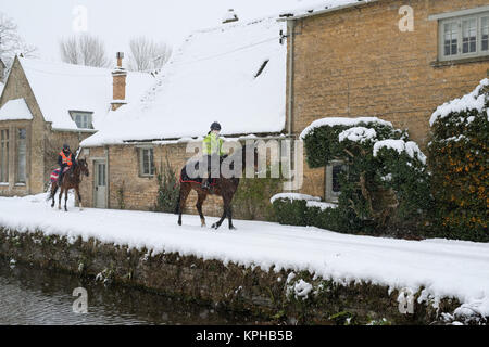 Les jeunes femmes de l'équitation à travers Lower Slaughter village dans la neige en décembre. Lower Slaughter, Cotswolds, Gloucestershire, Angleterre. Banque D'Images