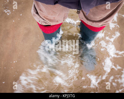 Homme portant des bottes wellington debout au bord de la mer avec l'eau floue autour de ses pieds Banque D'Images