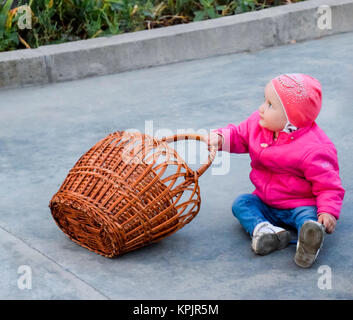 Un enfant dans un dossier rose et un chapeau s'assied sur le béton à côté d'un panier en osier Banque D'Images